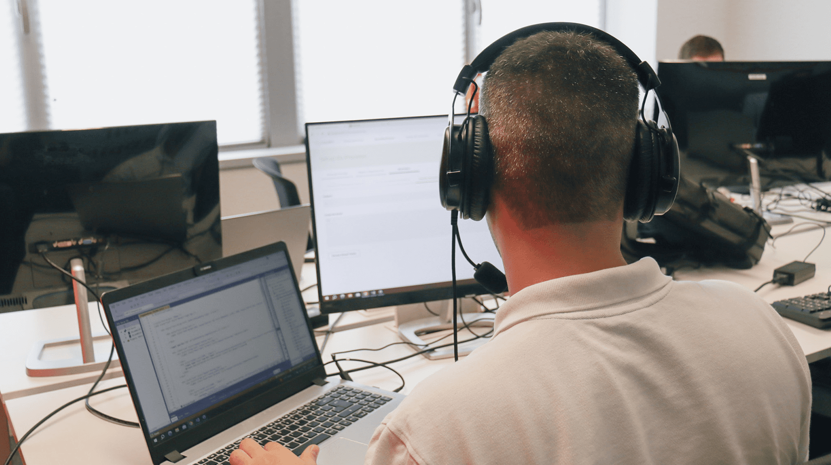 young man with his back turned, looking at his laptop and with an image on a second, larger computer screen. In the background we can see the office decoration in tones of white and several other computer screens