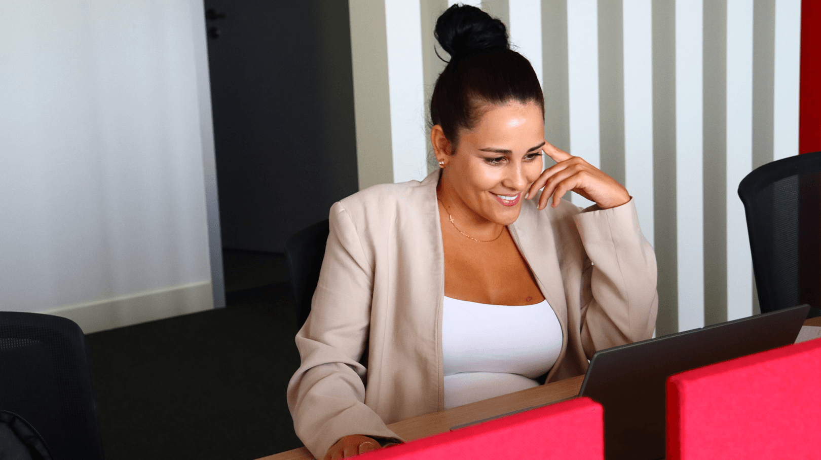 young brunette woman with ponytail, wearing a white top and a cream blazer, focused on her computer. In the background, the office in shades of white and red