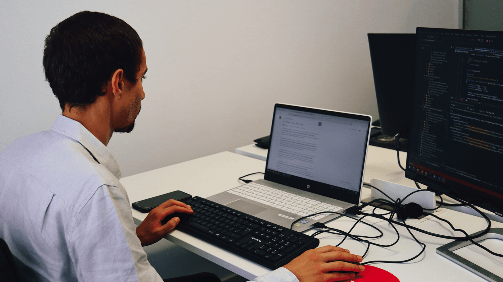 young man face to face working in the same office room, at the computer, programming. In the background we can see the white wall