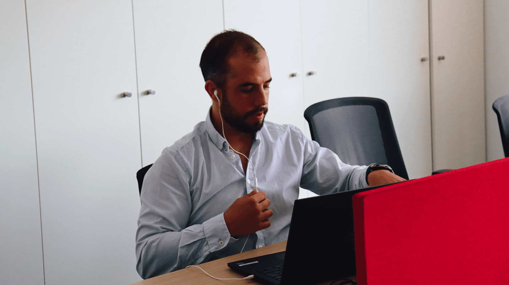 man focused and working on the computer, dressed in a baby blue shirt. In the background, a white cabinet and red decoration details