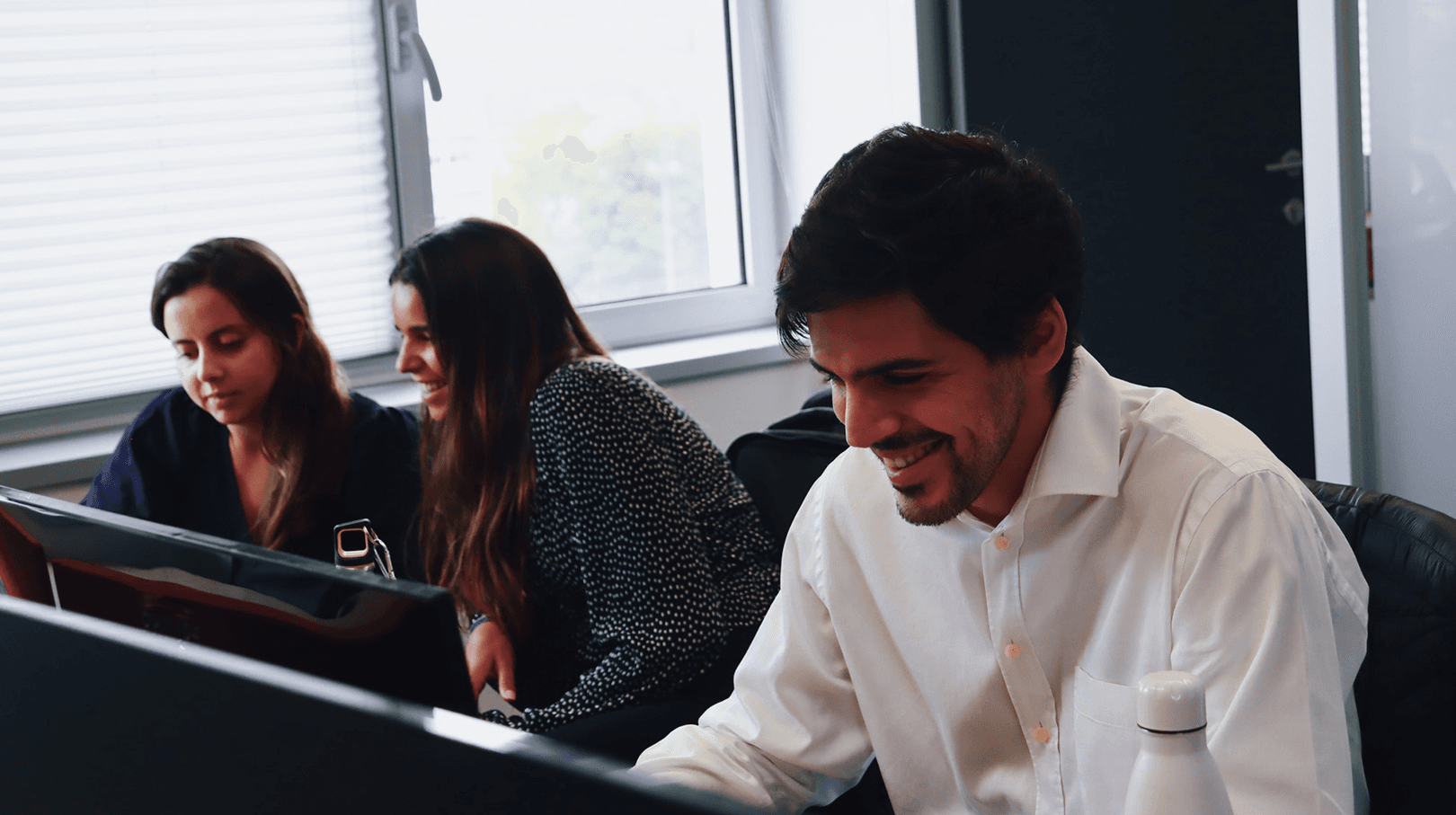 three red.it employees working together on the same work island. From left to right we can see a young brunette woman in dark blue, in the center another young brunette woman dressed in black, and on the right a young man in a white shirt. Everyone has laptops in front of them and all 3 of them are smiling when looking at them. In the background we can see details of the office in white, black and red.