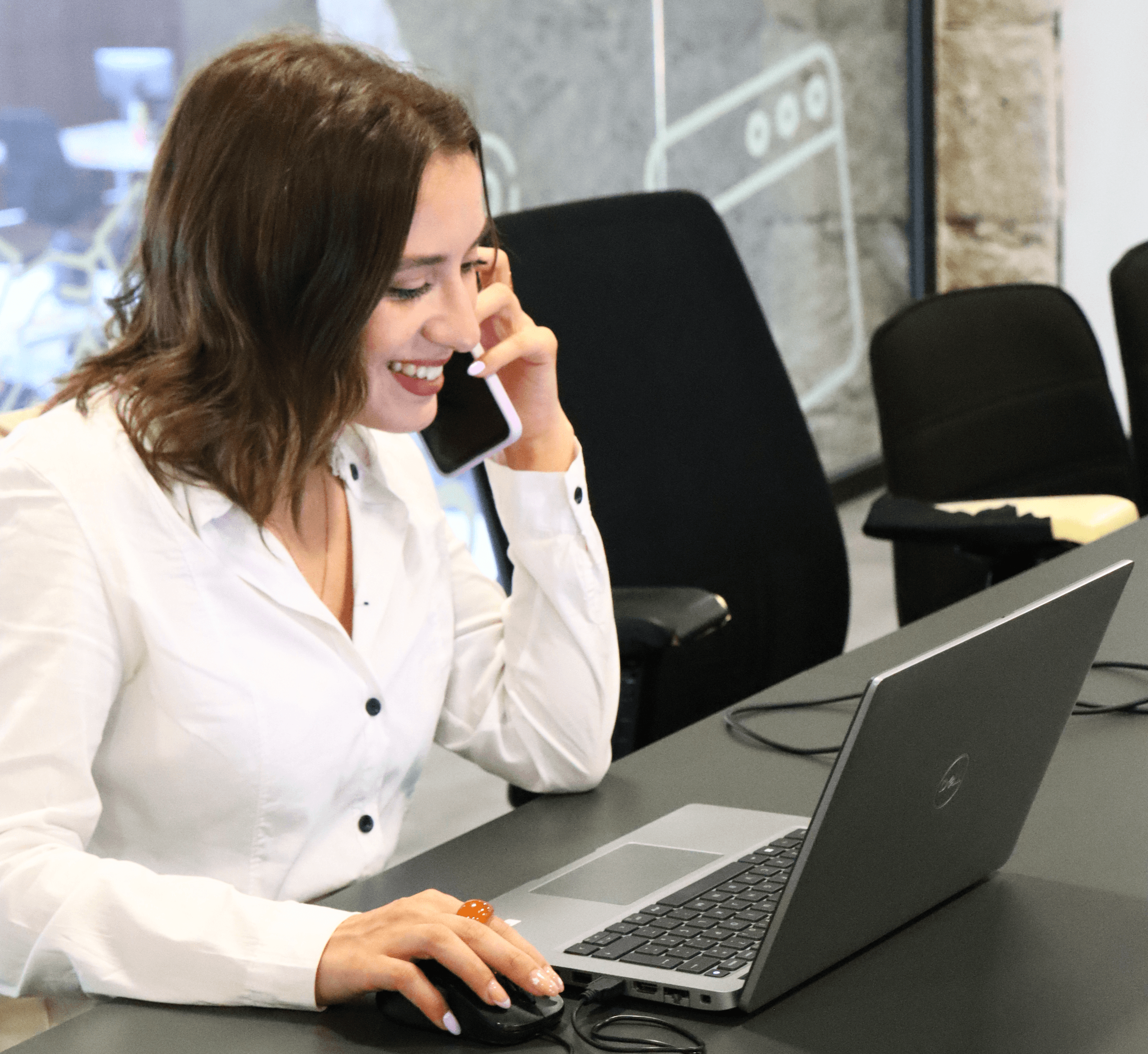 photograph of a young woman conducting a remote interview. Talking on the cell phone and with the computer in front of you. In the background, we can see a window that looks into the office.