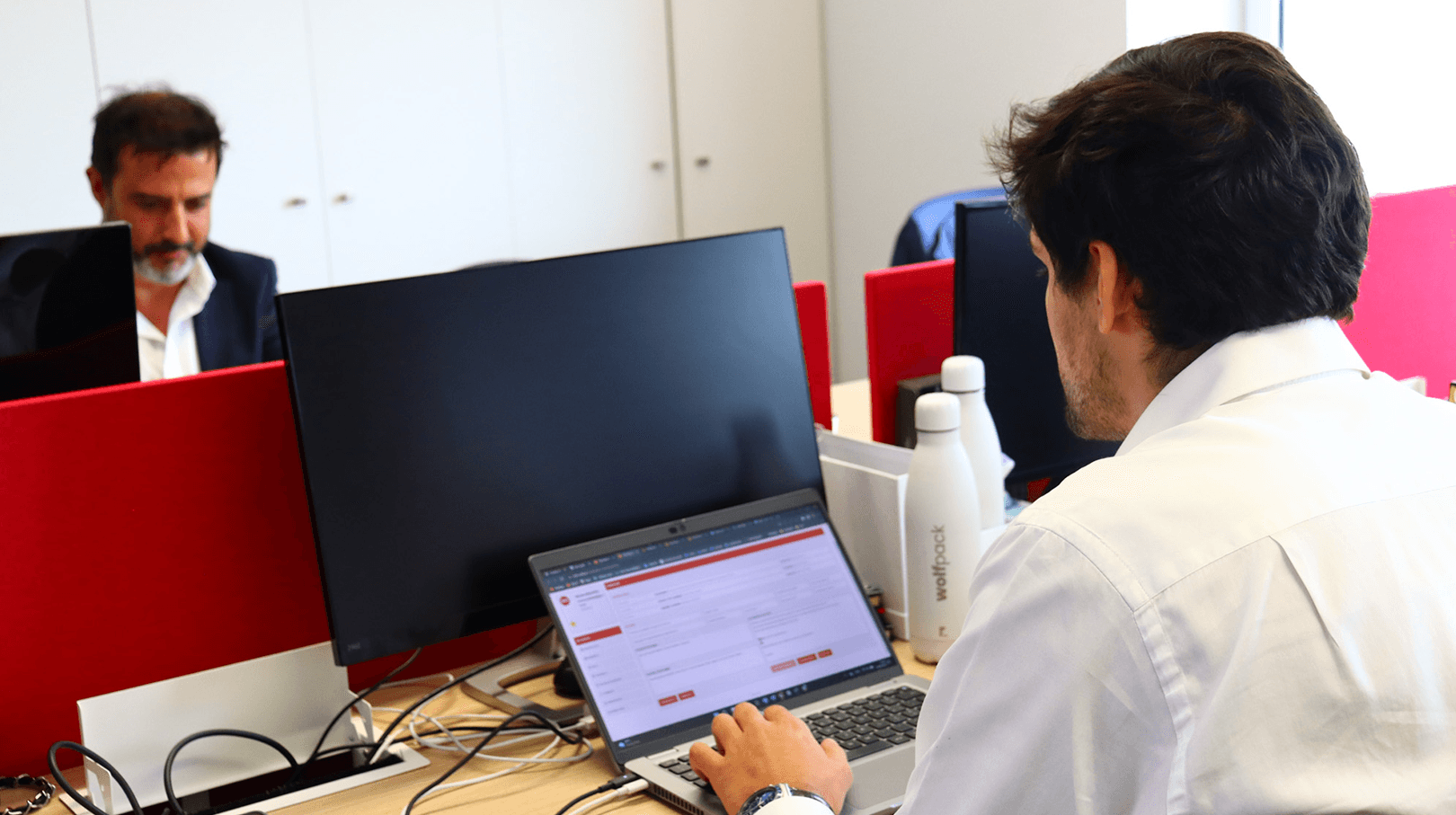 two co-workers working in front of each other at office desk, focused on their computer. Office background in red and white tones.