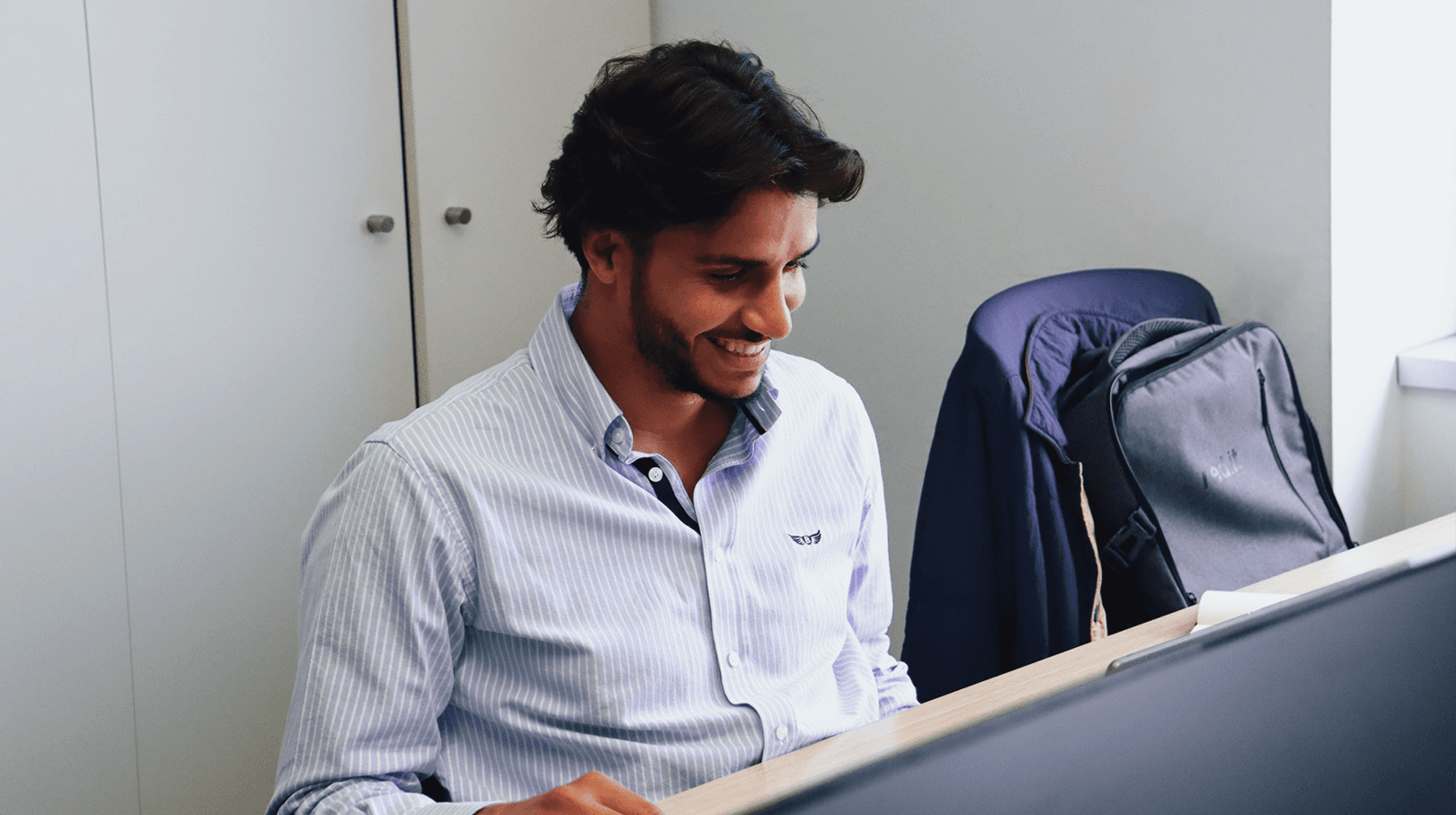 young man working on the computer with a smile, dressed in a white and baby blue striped shirt. On your left side there is another work chair with a gray red.it backpack. In the background we can see a white closet.