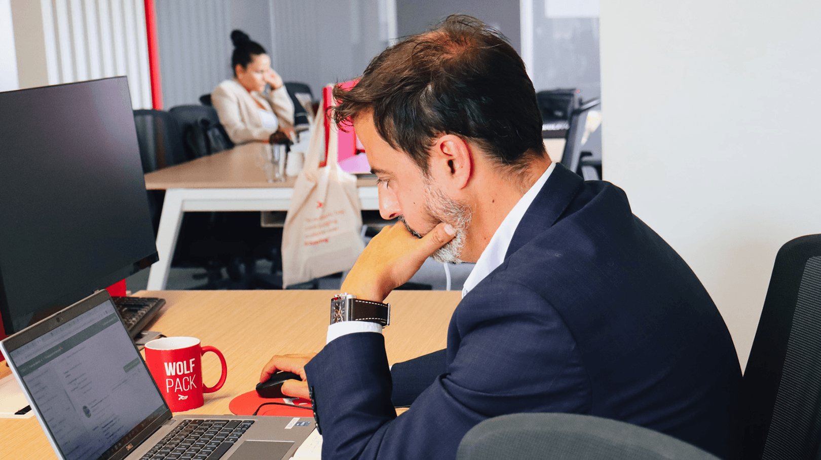 man focused on his work on the computer, looking concentrated and dressed in a blue suit and white shirt, with a watch on his left wrist. His desk at work has a red mug on top. In the background, we can see the office in shades of black, white and red.