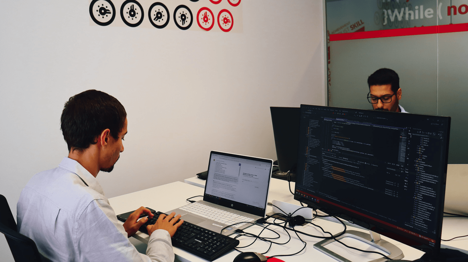 two young men face to face working in the same office room, at the computer, programming. In the background we can see the white wall with red and black elements.