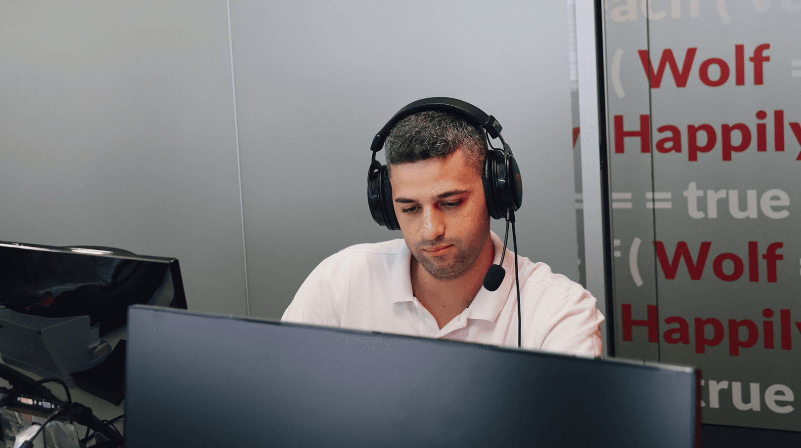 young man working in the office, at the computer. In the background we can see the gray wall with red and white elements associated with programming.