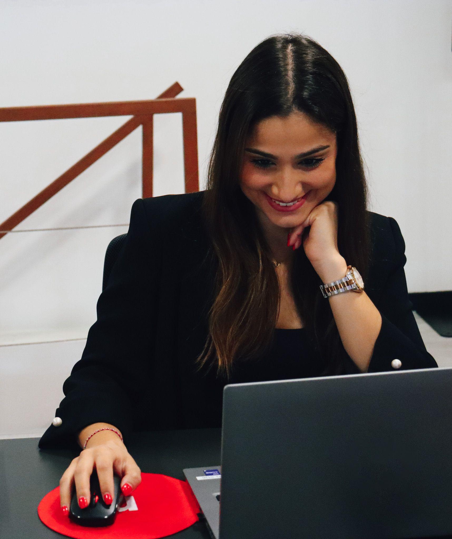 young woman working on the computer with a big smile, dressed in black and with red lipstick. In the background, we can see the office in shades of white and wood.