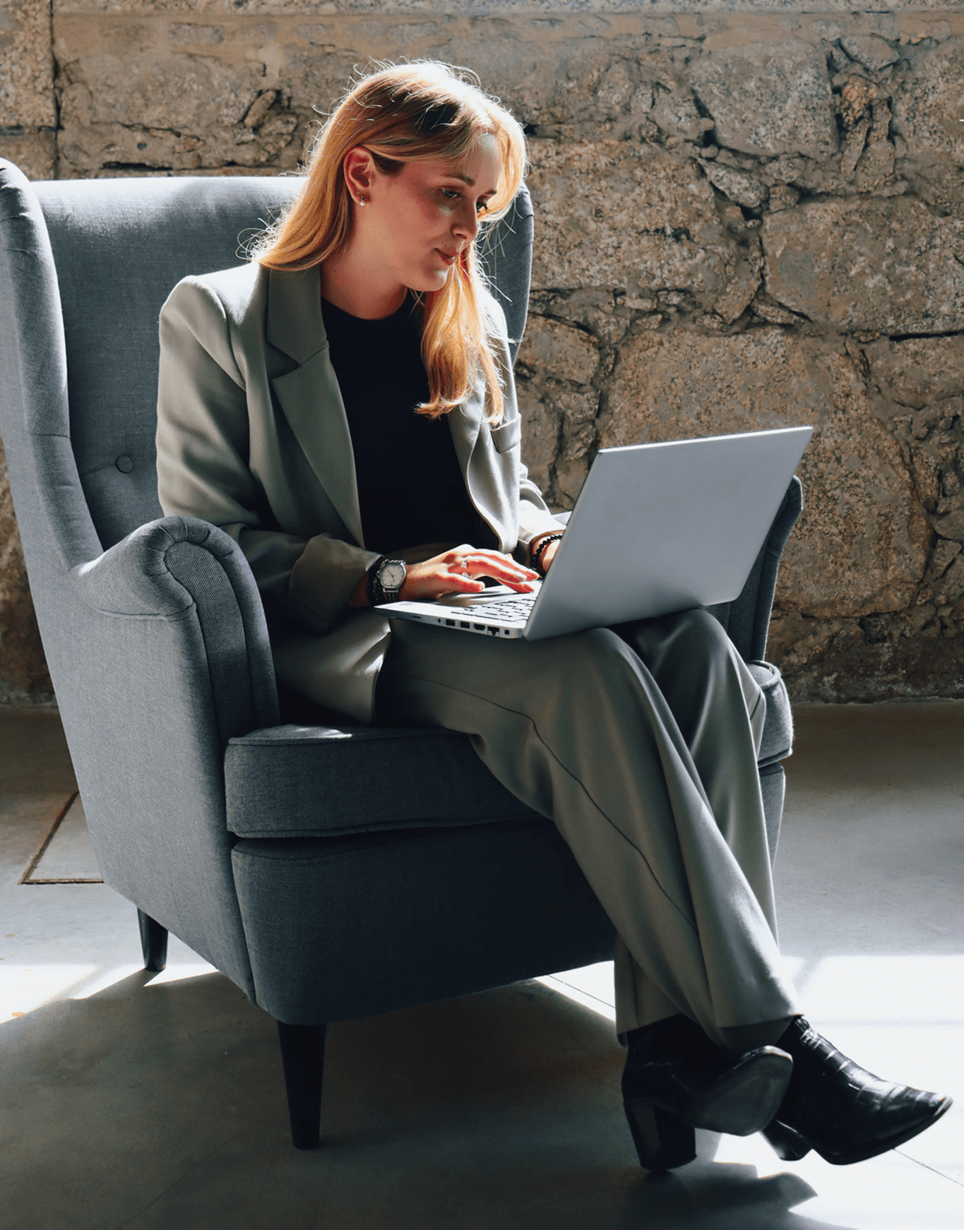 young woman working at her laptop sitting on a comfy chair. The backgound is a wall of stone.